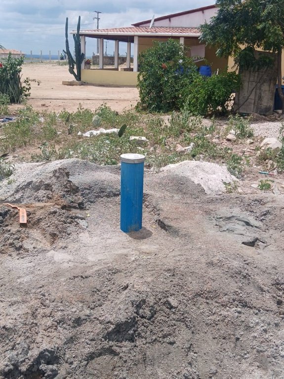 Tubo azul de poço artesiano recém-instalado e lacrado, cercado por terra removida em área rural com casa ao fundo.