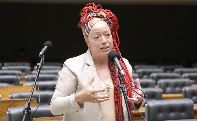 Deputada Daiana Santos fala ao microfone. Ela tem o cabelo vermelho, trançado e longo, usa um lenço colorido no cabelo e um blazer bege