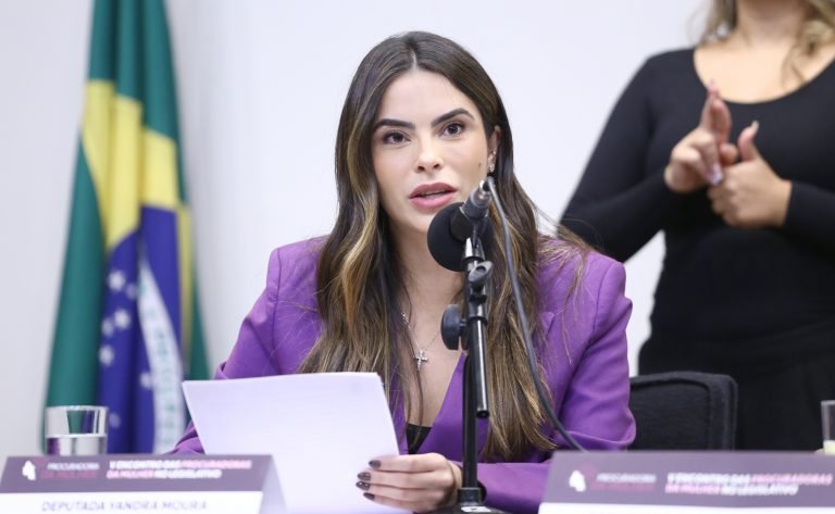 Deputada Yandra Moura fala ao microfone. Ela é uma mulher branca, tem cabelo longo, liso e castanho, veste um blazer roxo e segura um papel