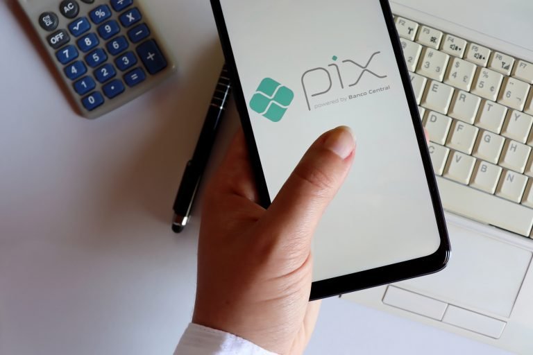 Uma mão segura um celular que exibe o logotipo do Pix, sistema de pagamentos do Banco Central. Ao redor, sobre uma mesa clara, há uma calculadora, uma caneta e parte do teclado de um computador.