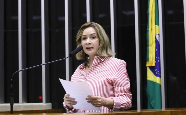 Deputada Socorro Neri fala ao microfone. Ela é uma mulher branca, tem o cabelo liso, loiro, na altura dos ombros, veste uma camisa listrada de vermelho e branco e segura um papel