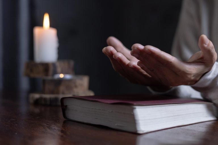 Mãos de uma pessoa abertas em gesto de oração sobre um livro fechado, apoiado em uma mesa de madeira. Ao fundo, à esquerda, há uma vela branca acesa.