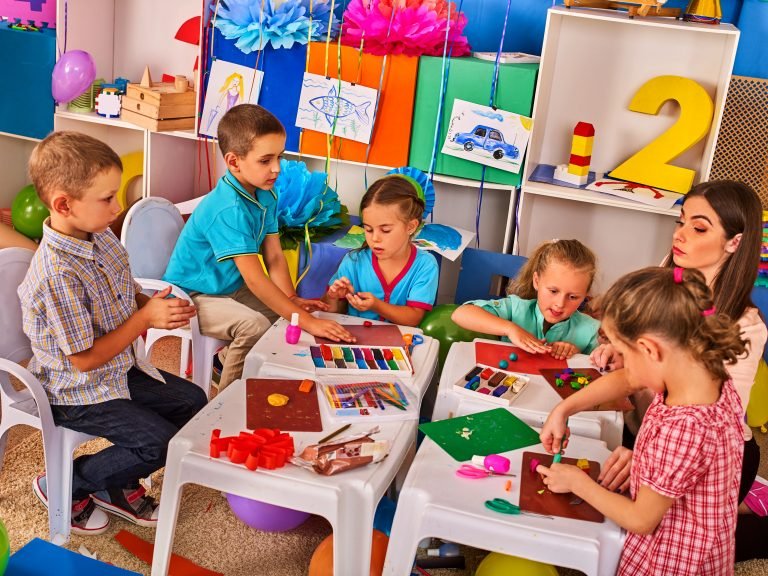 Crianças sentadas fazendo trabalhos manuais com papéis coloridos enquanto a professora observa