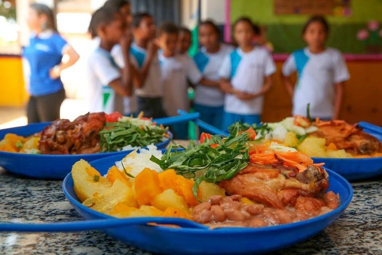 Em destaque na imagem, pratos com comida prontos para serem servidos. Ao fundo crianças, alunos de uma escola, aguardando para se alimentar.