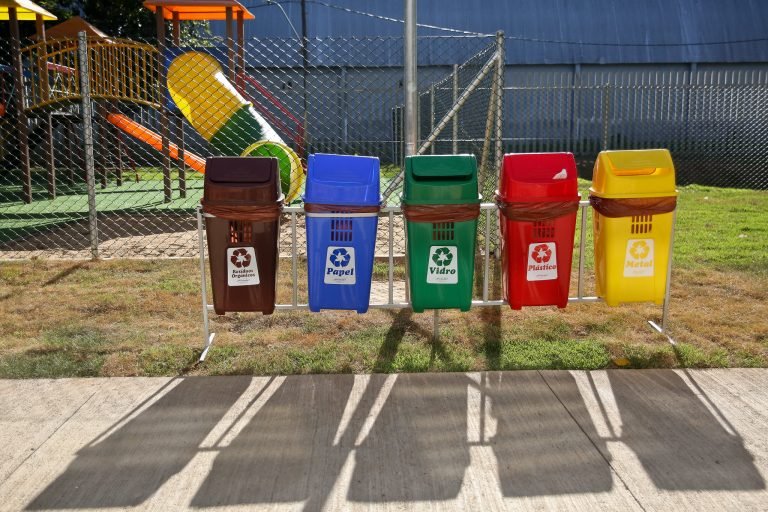Cinco lixeiras de coleta seletiva na seguinte ordem: marrom (resíduos orgânicos); azul (papel e papelão); verde (vidro); vermelha (plástico); e amarela (metal)